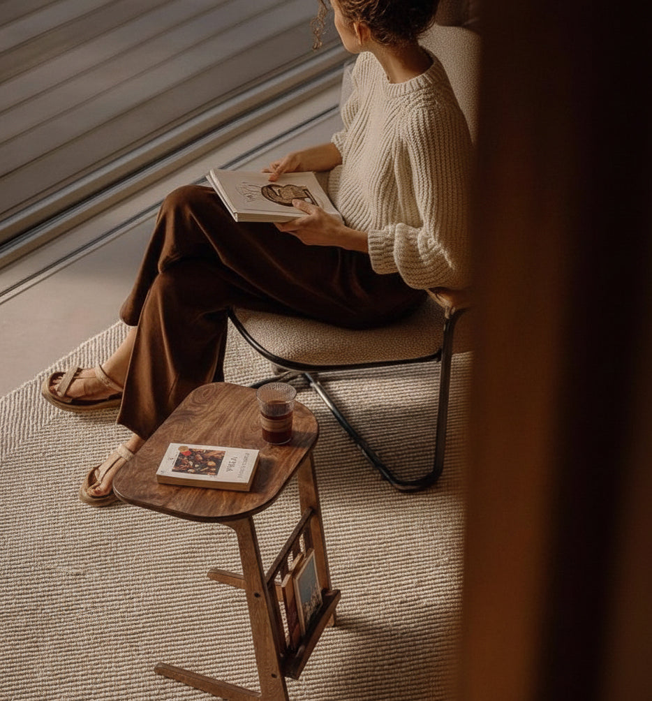 Pencere kenarında oturan bir kişinin yanında duran, üzerinde kahve ve kitap bulunan masif ahşap yan sehpa. Sehpanın alt kısmında dergilik bölmesi yer alıyor.